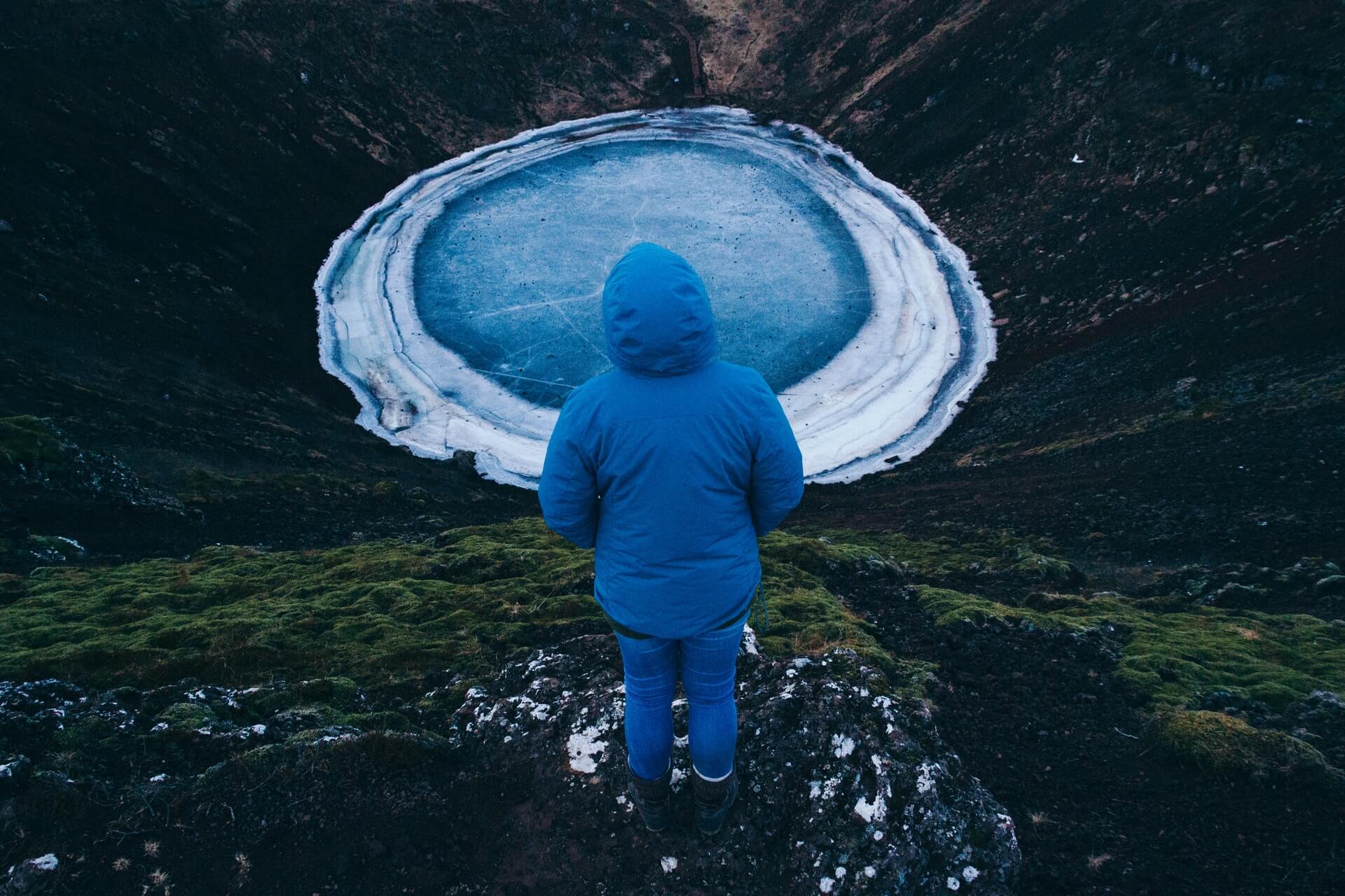 An aerial photograph of the Kerid Crater frozen in winter. The icy blue water stands out vividly against the dark volcanic rock. A person in a blue winter jacket stands with their back to the camera, overlooking the water. 